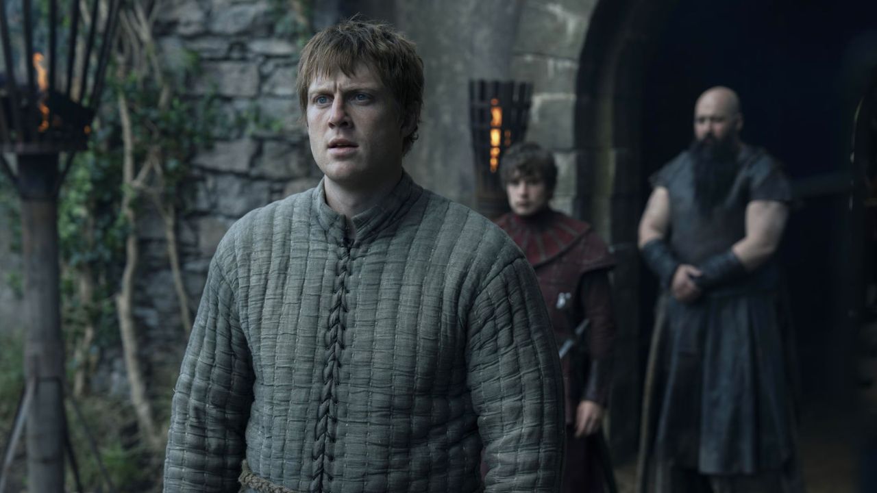 O Cavaleiro dos Sete Reinos Dunk em armadura diante da multidão durante o Julgamento dos Sete, com Baelor Targaryen ao fundo em clima tenso e sombrio.