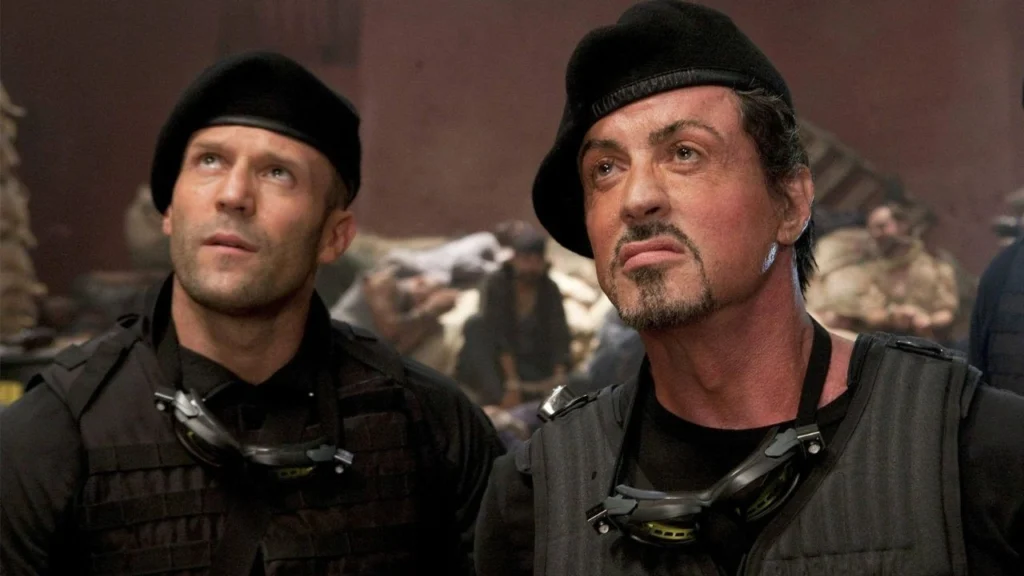 Jason Statham e Sylvester Stallone em cena de Os Mercenários 4, filme de ação marcado por tiroteios, explosões e clima militar.