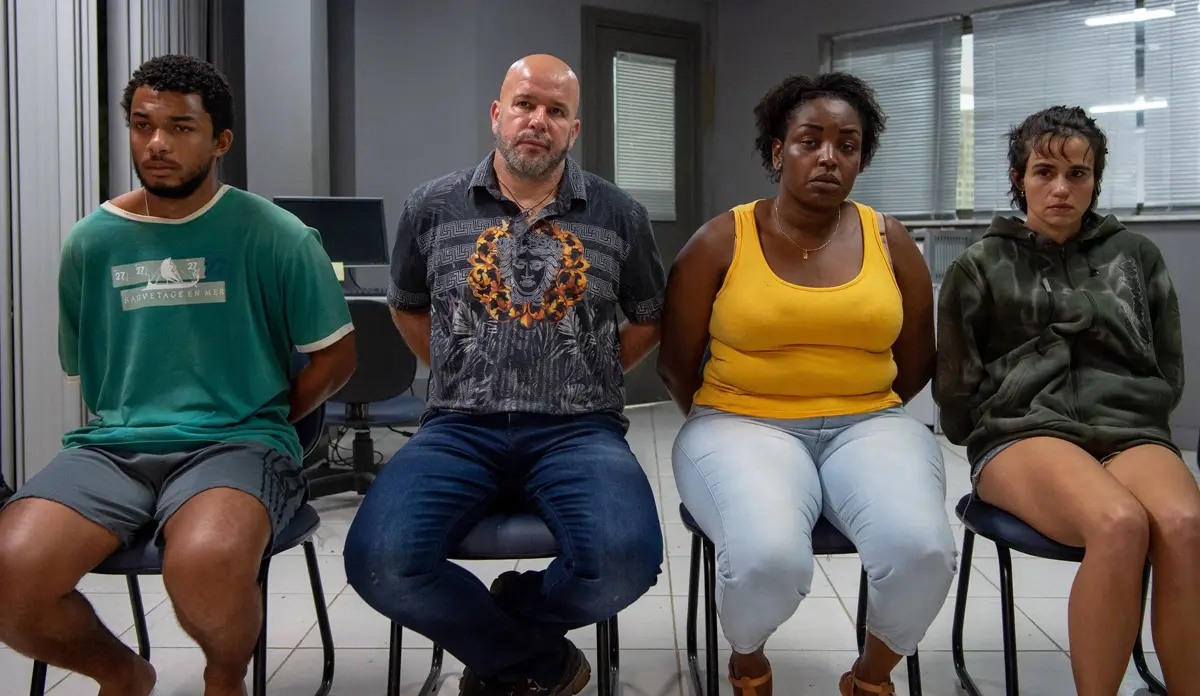 Cena de Justiça 2 mostra quatro personagens sentados em uma sala, com expressão séria.